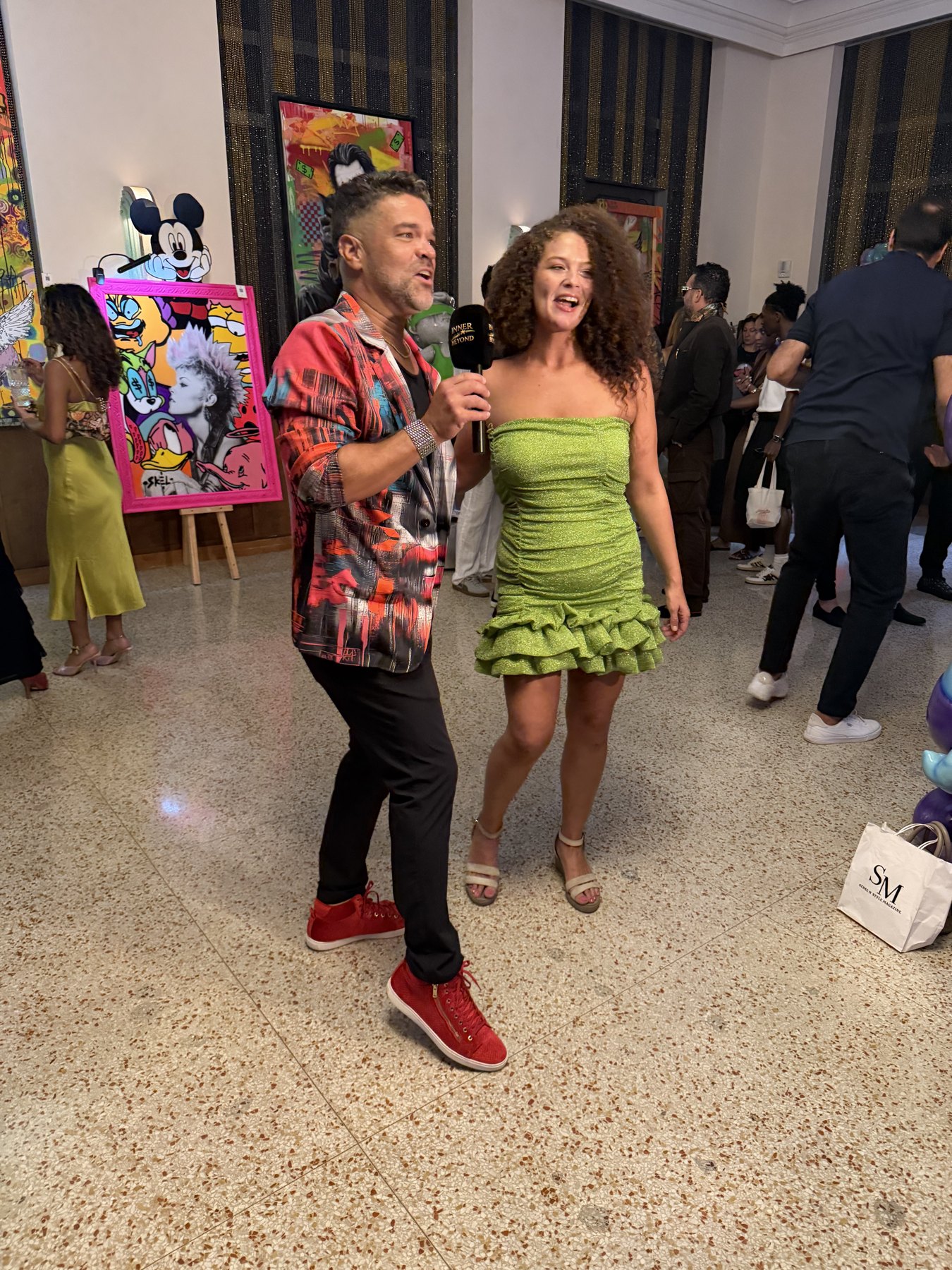 Jason Perez holding a microphone with a guest during a Jason Perez Art event surrounded by artwork